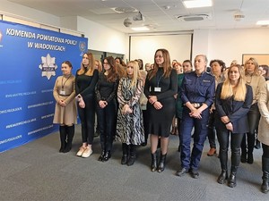 Grupa kobiet w strojach cywilnych oraz umundurowanych policjantek stojąca w szeregach. Po lewej stronie baner z napisem Komenda Powiatowa Policji w Wadowicach.